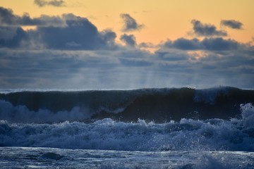 Storm on the sea at sunset. Blue sky with dark clouds. The light of the sun on the horizon. Big waves roll ashore. Splashes and mist over the wave.