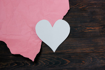 White paper heart lies on pink craft paper and wooden table. Blank for greeting card for Valentine's Day. Place for text
