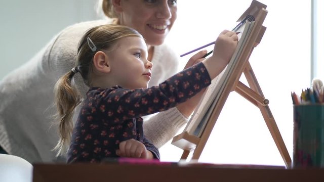 Mother showing adorable young daughter how to draw on easel at home