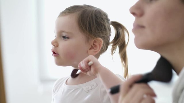 Adorable young daughter copying mother playing pretend with makeup brushes