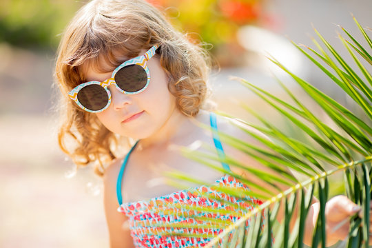 Closeup portrait of adorable little girl at the tropical resort, hiding behind the palm at the sunny summer day