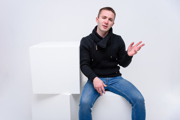 Concept studio portrait of a young man on a white background sitting on a cube and talking.