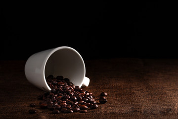 White cup full of coffee beans on the cloth sack in morning light and black background with copy space