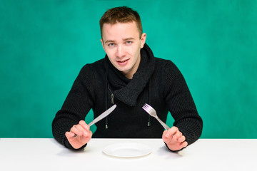 Concept young man eating at the table, holding a fork and spoon in hand, over a plate on a green background. He sits right in front of the camera with different emotions in different poses.