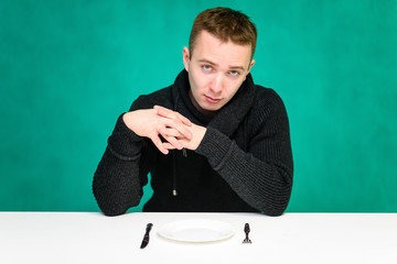 Concept young man eating at the table, holding a fork and spoon in hand, over a plate on a green background. He sits right in front of the camera with different emotions in different poses.