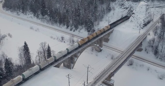 Freight train carries with gasoline tank an electric locomotive on railroad bridge by Trans Siberian two-sided railway along the river in the Ural Mountains - Aerial drone view