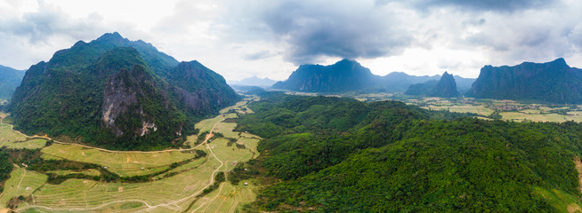 Naklejka na ściany i meble Aerial: Vang Vieng backpacker travel destination in Laos, Asia. Dramatic sky over scenic cliffs and rock pinnacles, rice paddies valley, stunning landscape.