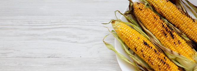 Grilled corn on the cob on a white plate over white wooden background, top view. From above,...