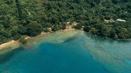 aerial drone image of a south pacific village on a remote island with a beautiful coral reef and lush tropical rainforest jungle while a small motor boat approaching