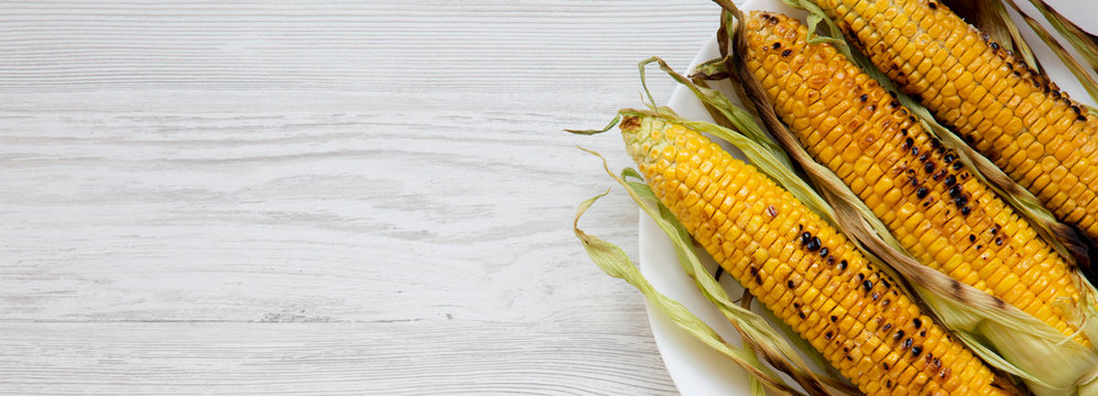 Grilled corn on the cob on a white plate over white wooden background, top view. From above, overhead. Copy space.