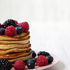 Pancakes with berries on a pink plate over white wooden background, side view. Copy space.