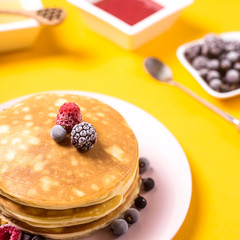 A stack of pancakes on a pink plate with berries next to honey and strawberry jam on a bright yellow background