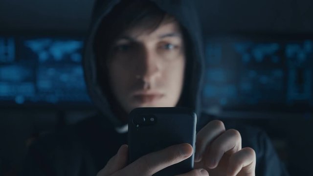 Dangerous Hacker in the hood uses a smartphone in a secret data center filled with screens