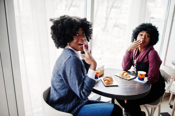 Two curly hair african american woman wear on sweaters sits at table cafe , eat croissant and drink tea.