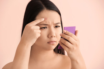 Portrait of young Asian woman with acne problem looking in mirror on color background