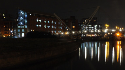 Fototapeta na wymiar a cityscape view of the canal entrance to the clarence dock area of leeds with a pedestrian bridge crossing the water with reflections of lights and buildings against a night sky with clouds