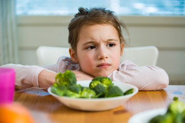 Cute girl eating spinach and broccoli at the table. Child doesn't want to eat, refuses eating, making faces. Healthy food concept. 