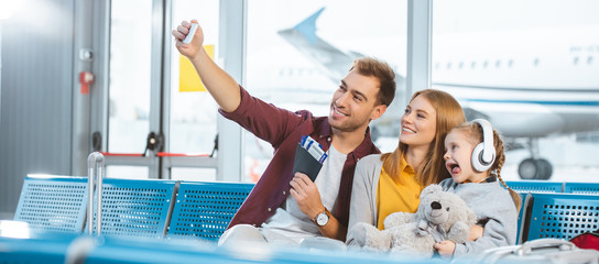 cheerful dad taking selfie and smiling with wife and daughter showing tongue in airport - obrazy, fototapety, plakaty