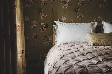 Interior view of bedroom with curtains and wallpaper with floral pattern, pale pink quilt and white pillows on double bed.