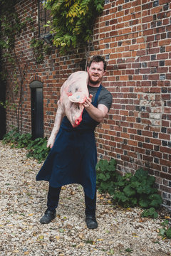 Male butcher wearing apron standing outdoors, carrying half a pig's carcass on his shoulder.