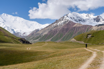 Man standing on tire tracks in a valley surrounded by snow capped mountains, Tulpar Kul, Kyrgyzstan, looking towards Peak Lenin.