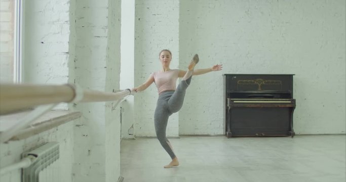 Graceful classic ballet dancer performing temps leve exersice at barre during rehearsal while training elements of classical dance in ballet studio. Ballerina practicing dance moves in dance studio