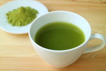 Closed Up a Cup of Hot Matcha Green Tea Served on Wooden Table with Blurry  Plate of Matcha Tea Powder in Background