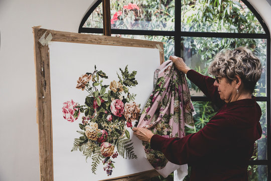 Senior woman wearing glasses and red dress standing in studio, comparing fabric pattern with painting of pink tea roses, leaves, berries and other flowers.