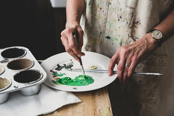 High angle close up of artist mixing green and brown colours on white plate.