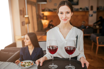 Nice young waitress stand and look straight on camera. She hold tray with two glasses of red wine. Customer sit behind at table and look to left. She has salad bowl on table.