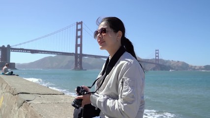 young girl travel photographer holding dslr camera sitting on rock happy smiling enjoy beautiful view by the bay. famous tourism attraction of san francisco golden gate bridge in background. - Powered by Adobe