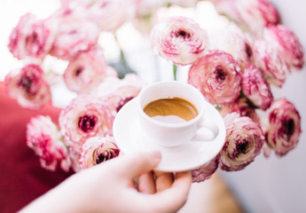 Hand holding a little cup of freshly brewed espresso coffee with a nice crema on the pink blossoming cappuccino Ranunculus flowers background, close up soft focus view