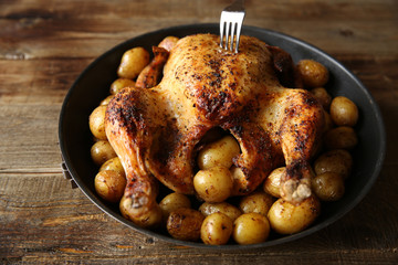 grilled chicken with a golden crust and golden potatoes in a frying pan with a fork in the chicken, decorated with green salad and red tomatoes on a wooden rustic background with space for text