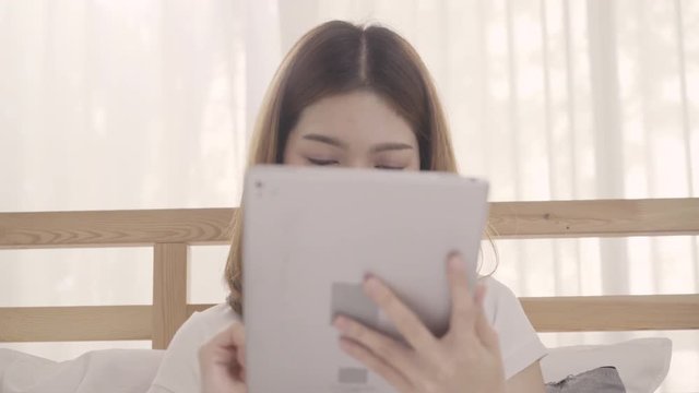 Young Asian woman using tablet while lying on bed after wake up in the morning, Beautiful attractive Japanese girl smiling relax in bedroom at home. Enjoying time lifestyle women at home concept.