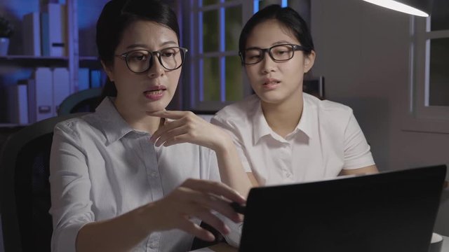 slow motion of two colleagues working overtime in dark office. Working a night shift in company using laptop computer discussing project hands pointing at screen monitor. young teamworker holding pen