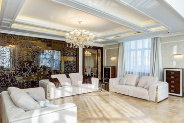 Classic interior of living room in brown, white, beige, with glasses wall and crystal chandelier in the center of ceiling. Three big and cozy sofas with pillows for big family.