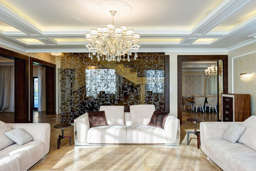 Big modern and luxurious interior of empty living room in brown, white, lithe brown colors, with pattern on glasses wall. Three sofas with big pillows, crystal chandelier in the center of ceiling.