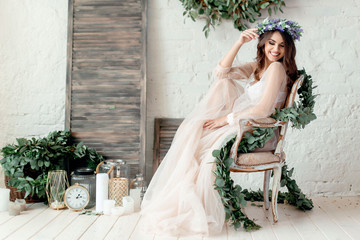 A girl in a beige peignoir, with a wreath of flowers on her head, poses in the studio loft, fine...