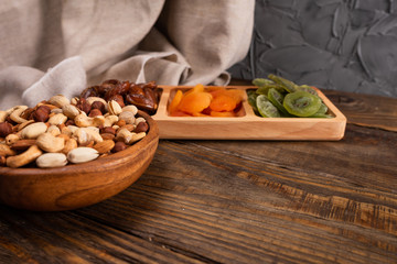 Dates, dried apricots and kiwis in a Compartmental dish and assortment of nuts in wooden bowl on a dark wooden table.
