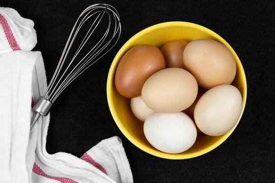 Top view of chicken eggs in a yellow bowl and an eggbeater on a black background