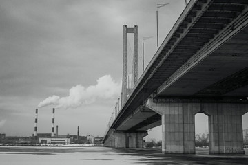 Bridge over the winter frozen river on the background of industrial smoking pipes. Cityscape