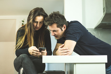 A young couple looking at the cell phone in the kitchen. Concept of technology