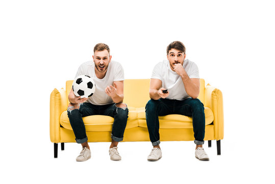 emotional young men with soccer ball sitting on couch and watching sports match isolated on white