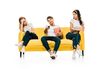 smiling young man with popcorn box and remote controller watching tv while bored and upset woman sitting on couch isolated on white
