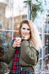 Portrait of white female model with bright makeup expressing positive energy.Portrait of cute girl with curvy curls smiling to camera in winter city. Holds a Cup of hot coffee or tea in his hands