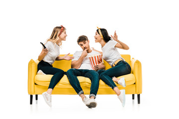 smiling young women talking and man eating popcorn while sitting together on sofa isolated on white