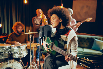 Obraz premium Mixed race woman singing and playing guitar while sitting on chair with legs crossed. In background drummer, saxophonist and bass guitarist.