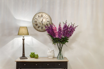 Crop of stylish and lighting corridor with wooden commode, retro watch over. Violet flowers in vase...
