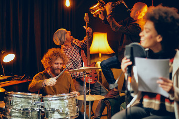 Mixed race woman singing. In background band playing instruments. Home studio interior.