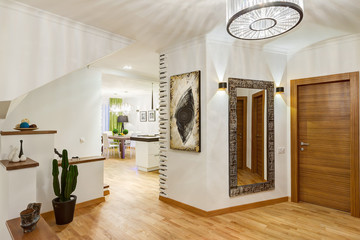 Modern corridor in big house in beige, white and brown colors. Stylish antechamber, with square mirror, picture on wall and chandelier in the center of the ceiling. Cactus in pots.
